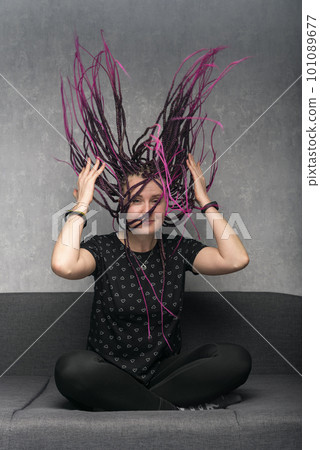 Portrait of happy young woman with long violet dreadlocks sitting on sofa. Girl waves her dreads. Vertical frame. 101089677