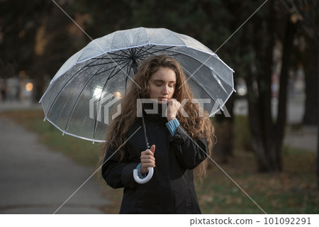 Brooding girl with long hair walks through autumn park in rainy weather under transparent umbrella. 101092291