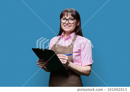 Middle aged woman in apron with clipboard posing on blue background Middle aged woman in apron with clipboard posing on blue background 101092813