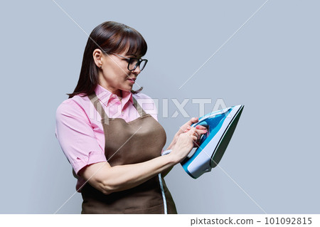 Woman in apron posing with iron, on gray background Woman in apron posing with iron, on gray background 101092815