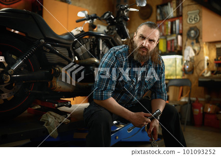 Portrait of mature bearded biker looking at camera sitting in his own garage 101093252