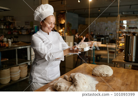 Woman baker in uniform sows flour through kitchen sieve on dough Woman baker in uniform sows flour through kitchen sieve on dough 101093399