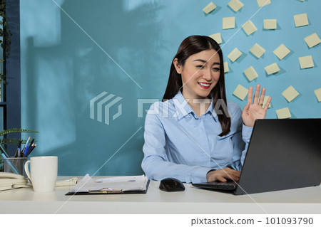 Corporate woman working from an office having a video call with work team with laptop. Female professional meeting virtually with business partners waving towards the screen. 101093790
