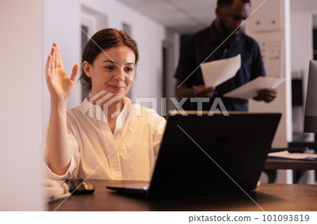 Smiling woman talking on videoconference, greeting colleague on videocall, waving hi. Corporate worker answering teleconference call in office at night, remote conversation in coworking space 101093819