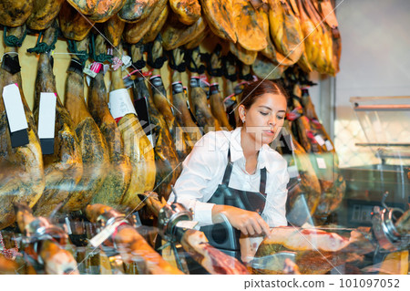 Young saleswoman in black apron slicing Iberian jamon at counter in butcher shop 101097052