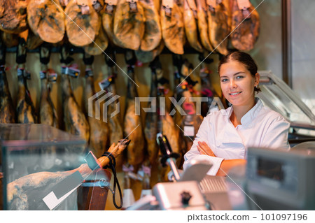 Young female butcher shopkeeper with crossed arms near hanging legs of Iberian ham Young female butcher shopkeeper with crossed arms near hanging legs of Iberian ham 101097196