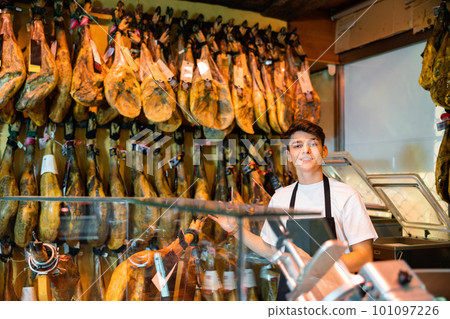 Young salesman in black apron offering dry-cured jamon in butcher shop Young salesman in black apron offering dry-cured jamon in butcher shop 101097226