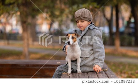 Caucasian boy sits on a bench with a dog Jack Russell Terrier for a walk in the autumn park.  101097238