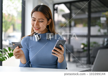 Attractive Asian woman sits at a table, holds her credit card and smartphone 101099144