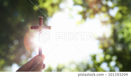 Fresh morning blue sky and green lush tree forest background and bright sun shining through the cross of Jesus Christ in hand 101103204