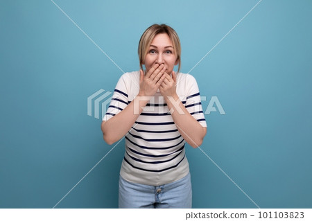 shocked blond young woman in a striped sweater covered her mouth with her hand in surprise on a blue 101103823