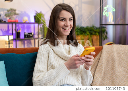 Young woman sitting on sofa at home, using smartphone share messages on social media application 101105743