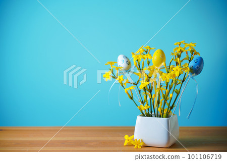 A bouquet of spring yellow flowers on blue background with easter eggs with pattern in a white vase 101106719