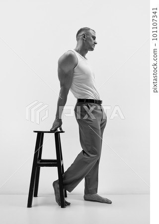 Portrait of elegant muscular man wearing pants and white t-shirt leaning on chair over studio background. Monochrome, side view 101107341