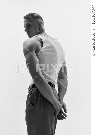 Portrait of muscular male fashion model wearing white t-shirt and posing over studio background. Monochrome, back view Portrait of muscular male fashion model wearing white t-shirt and posing over studio background. Monochrome, back view 101107344