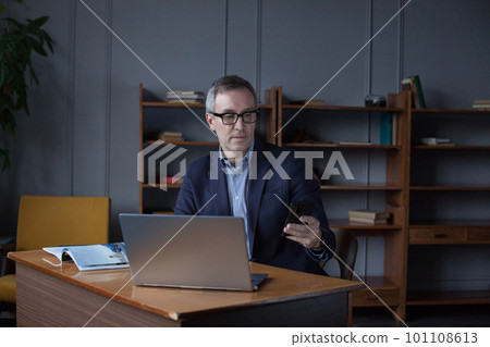 Portrait of mature attractive businessman in suit pensivly doing something on phone in his office Portrait of mature attractive businessman in suit pensivly doing something on phone in his office 101108613