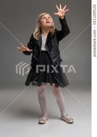 Little blonde girl in a white dress and black leather jacket is posing standing over a gray background. Little blonde girl in a white dress and black leather jacket is posing standing over a gray background. 101109520