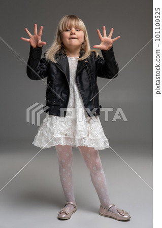 Little blonde girl in a white dress and black leather jacket is posing standing over a gray background. 101109525