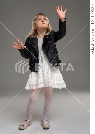 Little blonde girl in a white dress and black leather jacket is posing standing over a gray background. Little blonde girl in a white dress and black leather jacket is posing standing over a gray background. 101109526