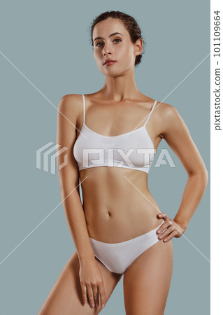 Young woman in white underwear, with bundled hair, hands on hips, posing against gray background. Plastic surgery, aesthetic cosmetology. Close-up. 101109664