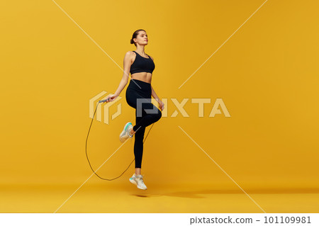 Sportive young girl with fit body training, jumping over jumping rope against yellow studio background. Cardio workout 101109981