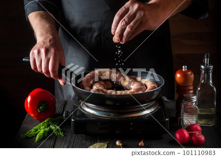 The chef adds salt to pan with raw meat sausage. Preparation for cooking sausages in the kitchen of a restaurant or cafe on table with vegetables and spices 101110983