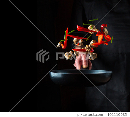Hand of a professional chef tosses pieces of vegetables and mushrooms on a frying pan on a black background. Restaurant cooking concept. Free advertising space Hand of a professional chef tosses pieces of vegetables and mushrooms on a frying pan on a black background. Restaurant cooking concept. Free advertising space 101110985