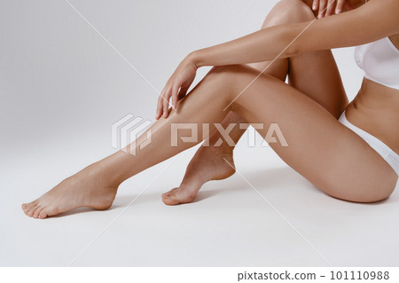 Perfect, slim, young body of a girl in white underwear sitting on the floor while posing sideways against grey background. Plastic surgery. Close-up. 101110988
