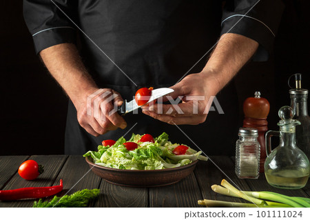 A professional chef prepares a vegetable salad in the restaurant kitchen. Cutting a fresh tomato for a vitamin salad with a knife. 101111024