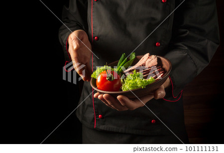Preparation of beef steak and fresh vegetables. Plate with food in the chef hand. Dark background free space for advertising. European cuisine. Preparation of beef steak and fresh vegetables. Plate with food in the chef hand. Dark background free space for advertising. European cuisine. 101111131