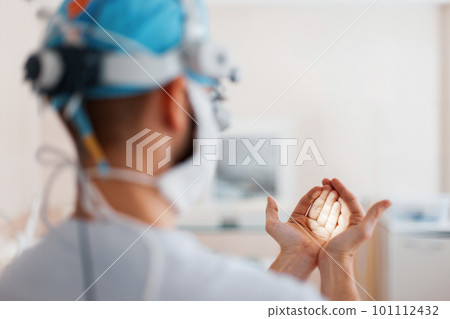 Doctor surgeon in medical mask in professional magnifying glasses shines on his hand with a flashlight, preparing for surgery. Back view Doctor surgeon in medical mask in professional magnifying glasses shines on his hand with a flashlight, preparing for surgery. Back view 101112432