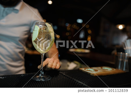 Glass with sweet white wine stands in the club on the bar counter against the background of the bartender in a white T-shirt. Tasty alcoholic drink for a good mood. Lifestyle Glass with sweet white wine stands in the club on the bar counter against the background of the bartender in a white T-shirt. Tasty alcoholic drink for a good mood. Lifestyle 101112839