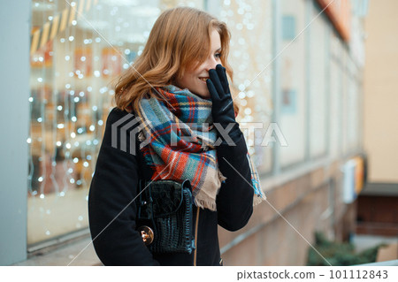 Positive young woman in a winter black coat in stylish gloves with a leather handbag with a woolen scarf is standing and laughing near the shop window decorated with festoons. Good mood. Cute girl. 101112843