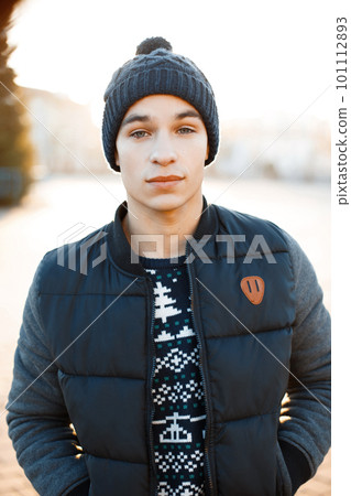 Cute charming young man in a blue stylish hat in a knitted vintage sweater in a fashionable warm jacket on winter sunny day American guy for a walk. 101112893
