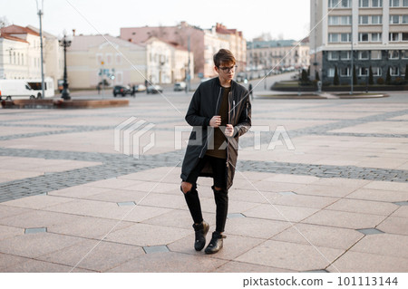 Handsome young hipster man in a fashionable autumn long jacket in torn stylish jeans in a t-shirt in trendy sneakers in stylish glasses posing in the city on the street. American modern guy. Handsome young hipster man in a fashionable autumn long jacket in torn stylish jeans in a t-shirt in trendy sneakers in stylish glasses posing in the city on the street. American modern guy. 101113144