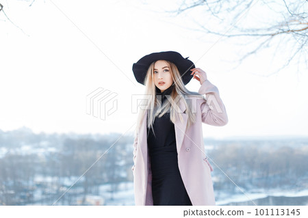Pretty cute beautiful young woman with blond hair in a vintage pink coat in a stylish hat in golf in a skirt is stands on the street in a winter sunny day. Fashionable girl walks outdoors. 101113145