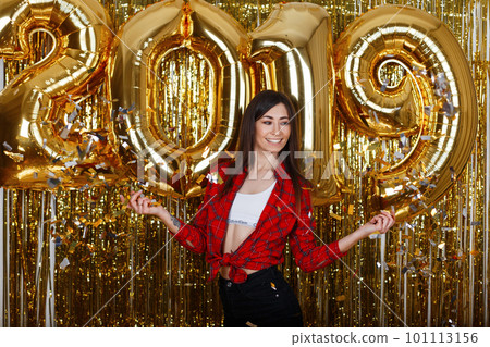 Cheerful young happy woman in fashionable clothes near the golden balloons of 2019 poses in the studio with flying confetti. Hurry to make a wish. Girl is having fun at the party. 101113156