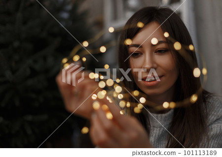 Young happy beautiful girl in a gray fashionable sweater with Christmas lights in her hands. Friendly girl smiling. Warm New Year mood. 101113189