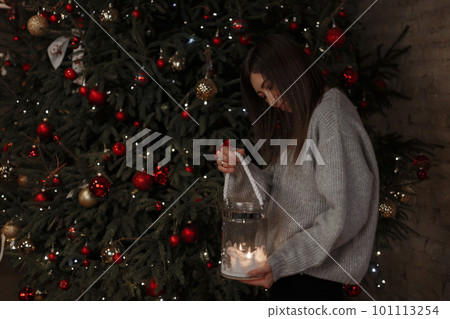 Young happy woman in a vintage knitted sweater is holding in hand a beautiful glass candlestick with a burning candle on the background of a Christmas tree decorated with toys. Cheerful girl. Young happy woman in a vintage knitted sweater is holding in hand a beautiful glass candlestick with a burning candle on the background of a Christmas tree decorated with toys. Cheerful girl. 101113254