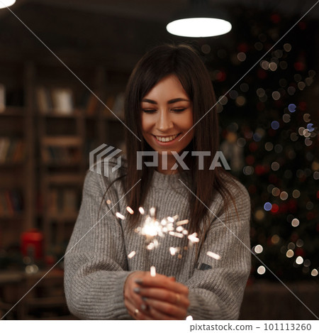 Young happy woman in a knitted sweater holds an amazing sparkler in her hand in a vintage room. Merry Christmas and Happy New Year. Girl smiling. 101113260
