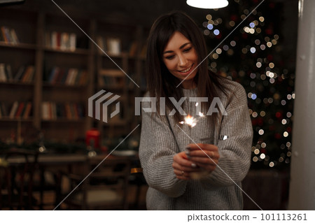 Cheerful young woman with a cute smile in a knitted sweater is standing and holding a sparkling sparkler in her hands near the background of the Christmas tree. Positive girl celebrates New Year. Cheerful young woman with a cute smile in a knitted sweater is standing and holding a sparkling sparkler in her hands near the background of the Christmas tree. Positive girl celebrates New Year. 101113261