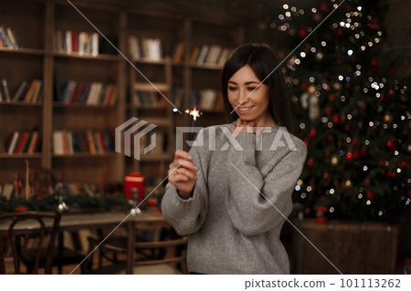 Young woman in a knitted warm sweater holds a sparkler in her hand smiles and looks at it near the background of a vintage bookcase in a festive room. Joyful girl. Young woman in a knitted warm sweater holds a sparkler in her hand smiles and looks at it near the background of a vintage bookcase in a festive room. Joyful girl. 101113262