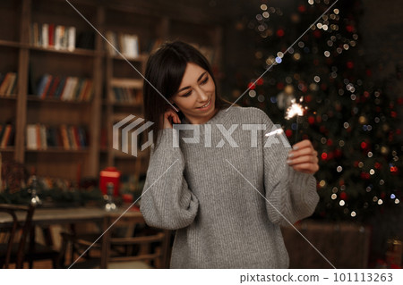 Beautiful young cheerful woman in a knitted fashionable sweater is holding a sparkler on the background of the Christmas tree in a vintage room. Magic New Year's atmosphere. Cute girl. 101113263