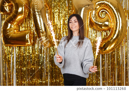 Positive young woman in a vintage knitted sweater with fireworks on the background of festive gold garlands and with New Year's air balloons 2019. Girl with a beautiful smile celebrates the New Year. 101113268