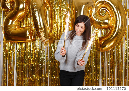 Happy young woman in a gray knitted sweater with fireworks on the background of golden garlands and New Year's balloons 2019. Girl with a beautiful smile is fun at the New Year's party. 101113270