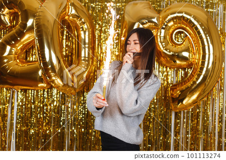 Happy young woman happily holds in her hands fireworks on the background of balloons 2019. The girl celebrates the New Year. Magical celebration atmosphere. Happy young woman happily holds in her hands fireworks on the background of balloons 2019. The girl celebrates the New Year. Magical celebration atmosphere. 101113274
