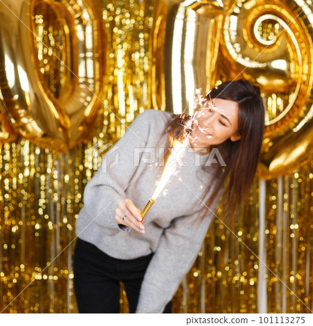 Young pretty woman in knitted sweater enjoy with fireworks in hands near the background of golden balloons 2019 and garlands in a festive studio. Girl is fun at the New Year's party. 101113275