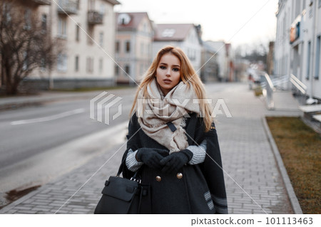 Fashionable attractive young blond woman in a vintage knitted sweater in a stylish coat in warm gloves with a vintage scarf with a fashionable black leather bag walking on the street. City cute girl. Fashionable attractive young blond woman in a vintage knitted sweater in a stylish coat in warm gloves with a vintage scarf with a fashionable black leather bag walking on the street. City cute girl. 101113463