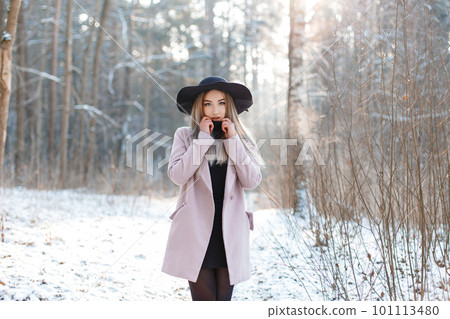 Young stylish blonde woman in an elegant black hat in a pink stylish coat in a black fashionable dress is standing and looking at the camera in the winter forest. Beautiful girl enjoy the scenery. Young stylish blonde woman in an elegant black hat in a pink stylish coat in a black fashionable dress is standing and looking at the camera in the winter forest. Beautiful girl enjoy the scenery. 101113480