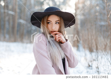 Amazing glamorous pretty beautiful young woman in a luxurious black hat in a stylish pink warm coat posing on a sunny day in a winter park. Sexy fashionable girl on a walk. Amazing glamorous pretty beautiful young woman in a luxurious black hat in a stylish pink warm coat posing on a sunny day in a winter park. Sexy fashionable girl on a walk. 101113481